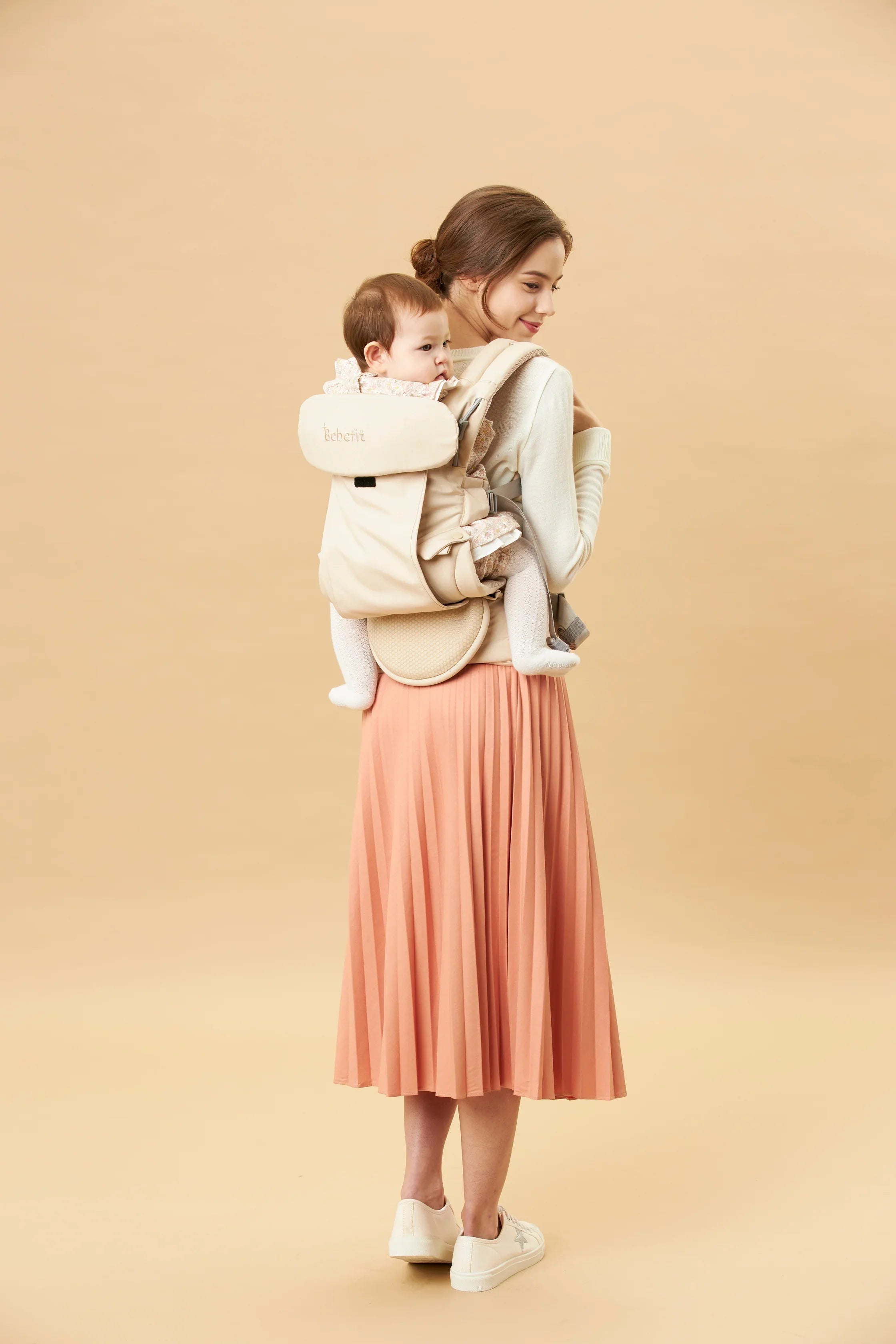 Mother carrying baby in beige bebefit.eu ergonomic baby carrier, modern neutral background